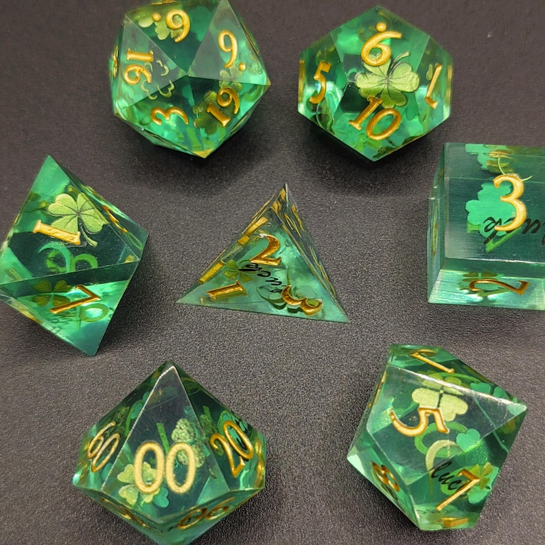 K&ouml;nnte beinhalten: Ein Set aus sieben gr&uuml;n-goldenen polyedrischen W&uuml;rfeln. Jeder W&uuml;rfel ist transparent mit goldenen Zahlen und Kleeblatt-Designs. Die W&uuml;rfel sind auf einer dunklen Oberfl&auml;che angeordnet, um ihre vielf&auml;ltigen Formen zu zeigen.