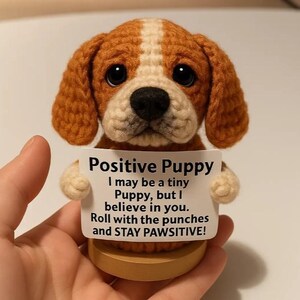 Könnte beinhalten: Gehäkelte Welpenfigur mit braun-weißem Fell, die ein Schild mit der Aufschrift "Positive Puppy" hält. Das Schild enthält den Text "I may be a tiny Puppy, but I believe in you. Roll with the punches and STAY PAWSITIVE!"