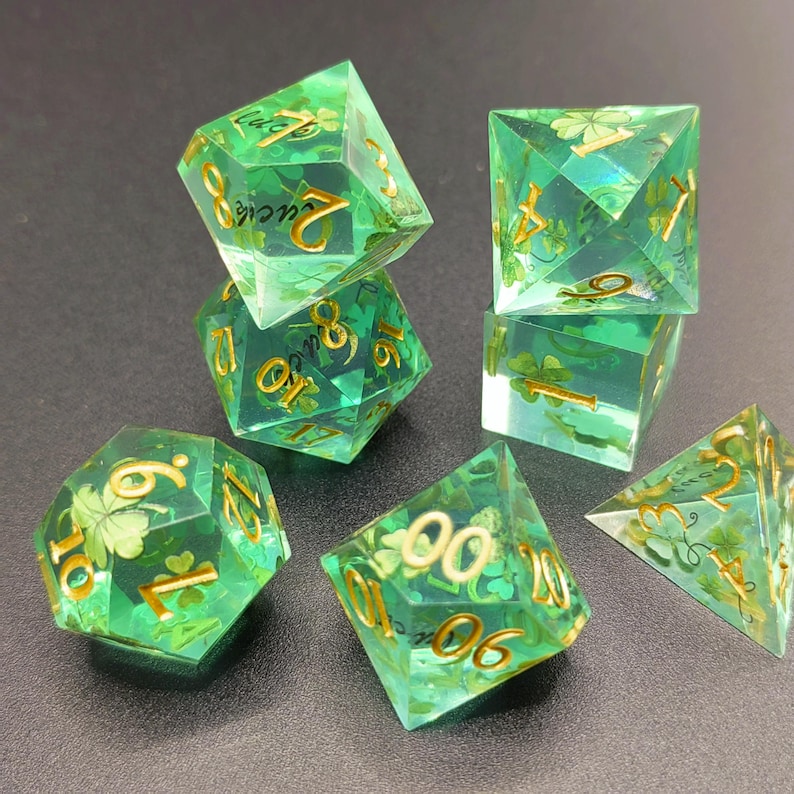 K&ouml;nnte beinhalten: Set aus sieben gr&uuml;nen Kunstharz-W&uuml;rfeln mit goldfarbenen Zahlen und Kleeblatt-Designs. Die W&uuml;rfel sind mehrseitig, darunter ein d4, d6, d8, d10, d12, d20 und ein Prozentw&uuml;rfel. Die W&uuml;rfel sind transparent.