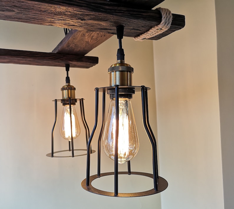 May include: Two black caged pendant lights with gold accents hang from a rustic wooden beam. The lights are turned on and illuminate the room with a warm glow.