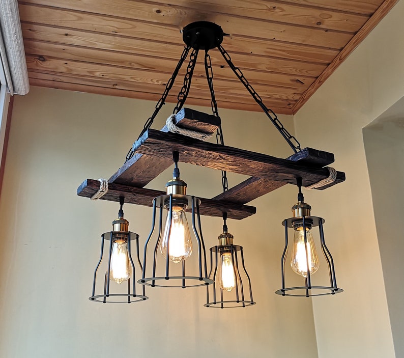 May include: A rustic wooden chandelier with five black metal cage light fixtures. The chandelier is suspended from the ceiling by chains and has a rope detail.