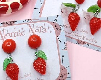May include: Two pairs of strawberry-shaped dangle earrings with red spherical studs, green leaves, and white flower accents, displayed on branded Atomic Nora cards alongside two cream-coloured bangles with red polka dots.