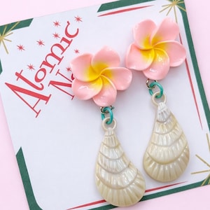 Può includere: Un paio di orecchini pendenti con perni a forma di fiore di plumeria rosa e giallo, collegati a ciondoli a forma di conchiglia color crema tramite piccoli anelli turchesi. Gli orecchini sono esposti su un cartoncino con il testo Atomic Na.