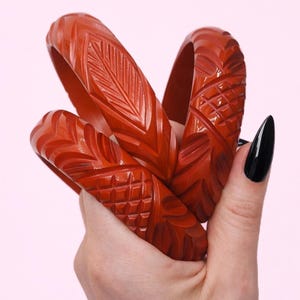 May include: Three deep red bangles featuring intricate carved patterns. The bracelets display a combination of textured leaf-like motifs and geometric grid designs across their surfaces. These circular accessories have a smooth, polished finish.