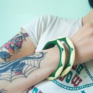 May include: Two circular bangle bracelets featuring a repeating pattern of alternating cream-colored and dark green segments. The bracelets have a smooth, polished finish and a rounded, thick profile, worn together on the forearm.