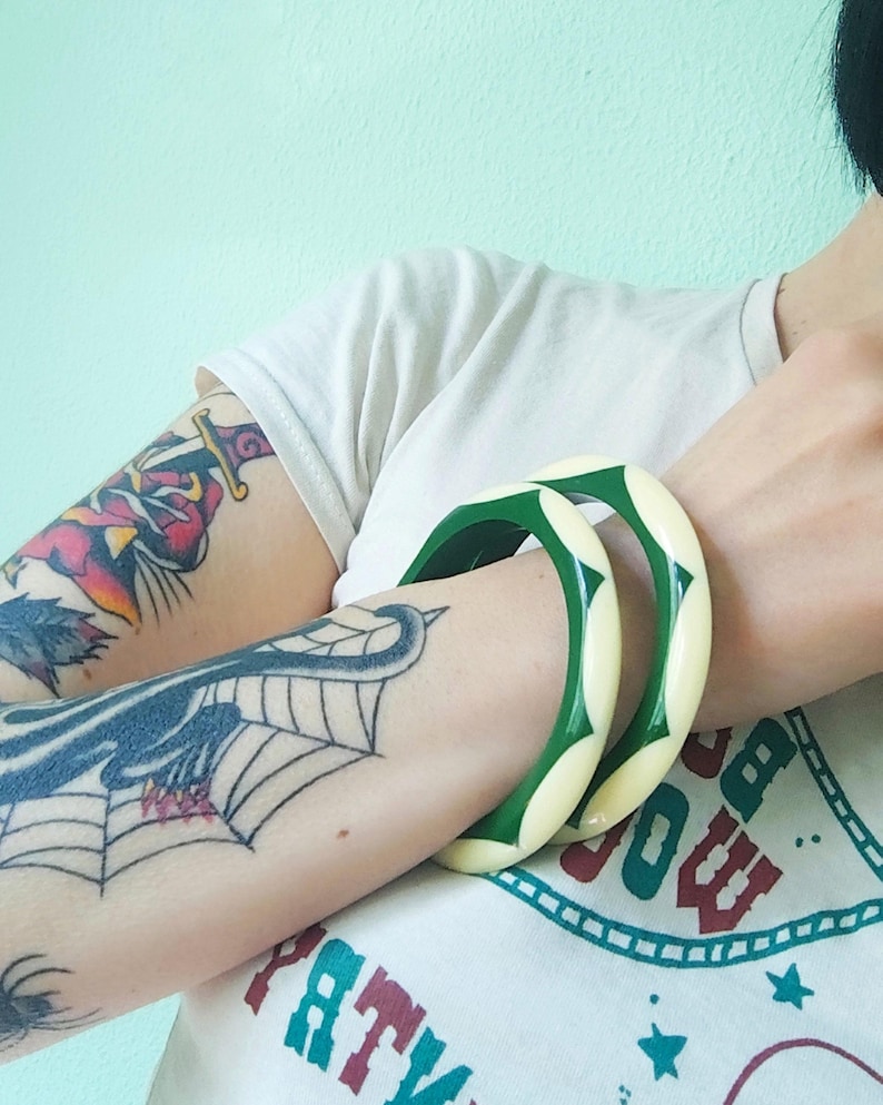 May include: Two circular bangle bracelets featuring a repeating pattern of alternating cream-colored and dark green segments. The bracelets have a smooth, polished finish and a rounded, thick profile, worn together on the forearm.
