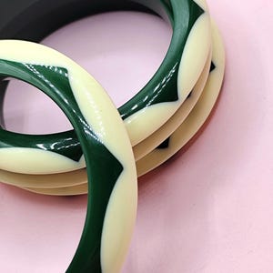 May include: A set of three circular bangle bracelets featuring a smooth, polished finish. Each bangle displays a repeating geometric pattern of alternating dark forest green and cream-colored segments arranged in a continuous, interlocking design.
