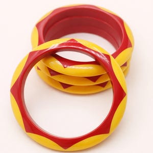 Peut inclure: Un ensemble de trois bracelets joncs circulaires présentant une base jaune vif avec un motif triangulaire rouge répétitif sur toute la circonférence extérieure. Les bracelets ont une finition lisse et brillante et sont empilés.
