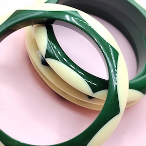 May include: Two circular bangle bracelets featuring a smooth, glossy finish. The design consists of alternating geometric segments in deep forest green and off-white cream colors, creating a repeating pattern around the circumference of each bangle.