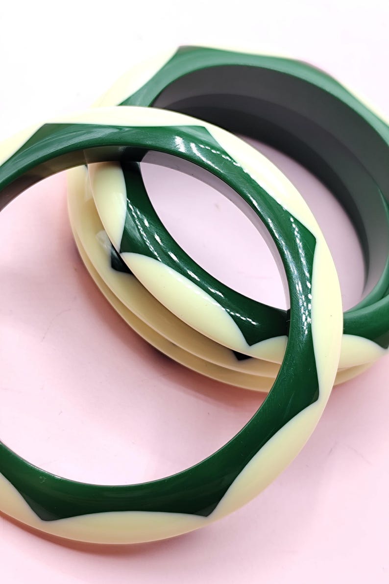 May include: Two circular bangle bracelets featuring a smooth, glossy finish. The design consists of alternating geometric segments in deep forest green and off-white cream colors, creating a repeating pattern around the circumference of each bangle.