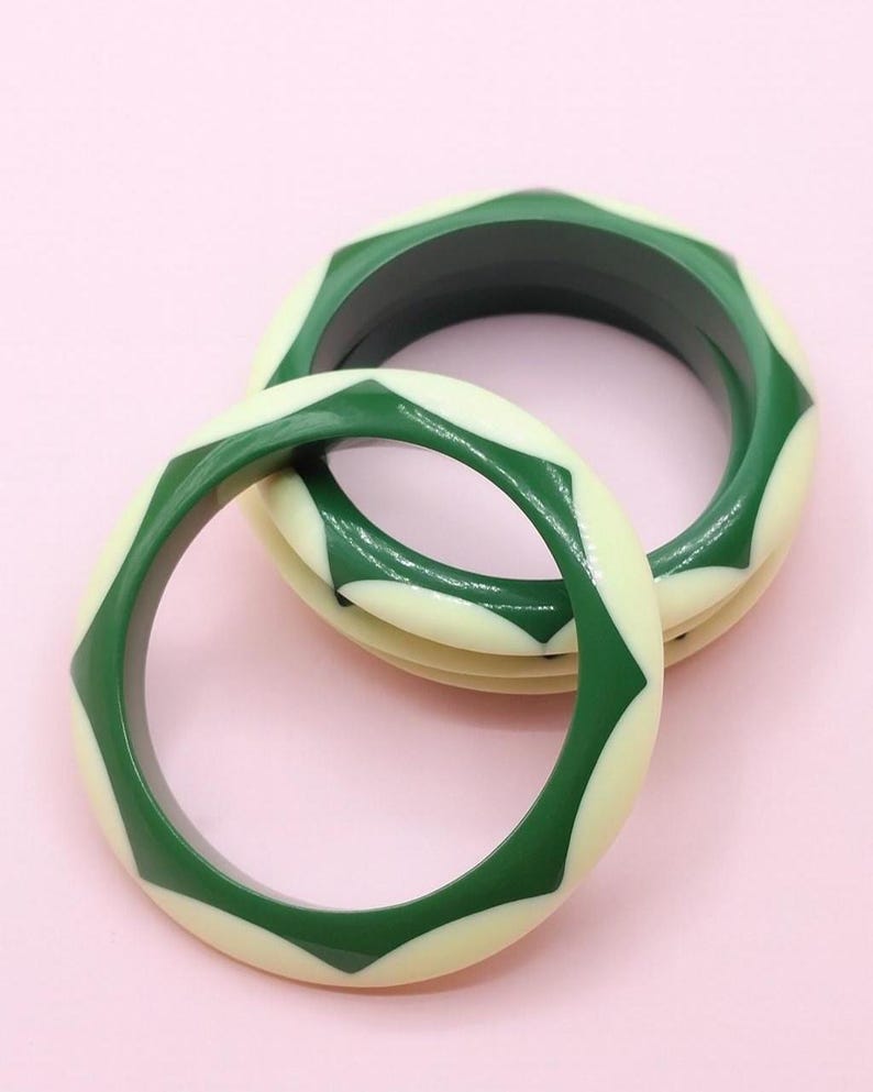 Può includere: Pila di braccialetti geometrici verdi e color crema. I braccialetti presentano un motivo ripetuto di forme a diamante verdi su sfondo crema. I braccialetti sono impilati su uno sfondo rosa.