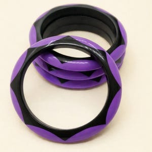 Pode incluir: Um conjunto de três pulseiras circulares rígidas com um padrão repetitivo de formas triangulares alternadas em roxo e preto. As pulseiras têm um acabamento liso e brilhante e estão empilhadas, exibindo o design geométrico.