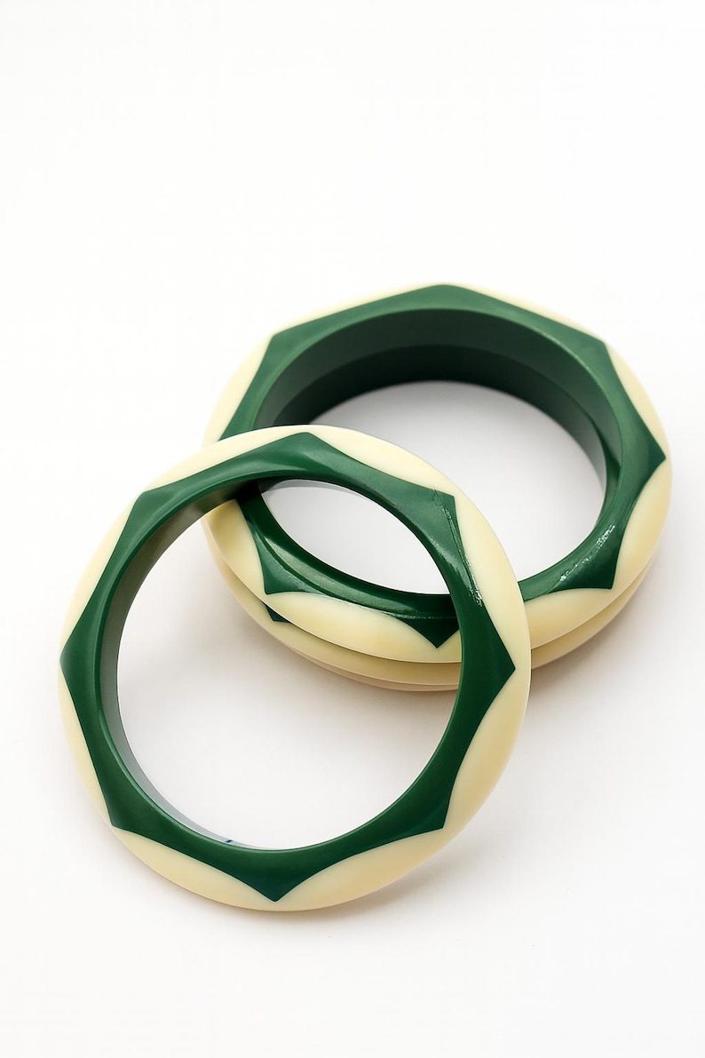 May include: Two circular bangles featuring a smooth, glossy finish. Each bangle displays an alternating geometric pattern of dark forest green and cream-colored segments, creating a repeating star-like design around the entire circumference.
