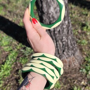 May include: A set of five circular bangle bracelets featuring a repeating pattern of cream-colored and dark green geometric shapes. The bracelets have a smooth, rounded surface and are designed to be worn stacked together on the wrist.