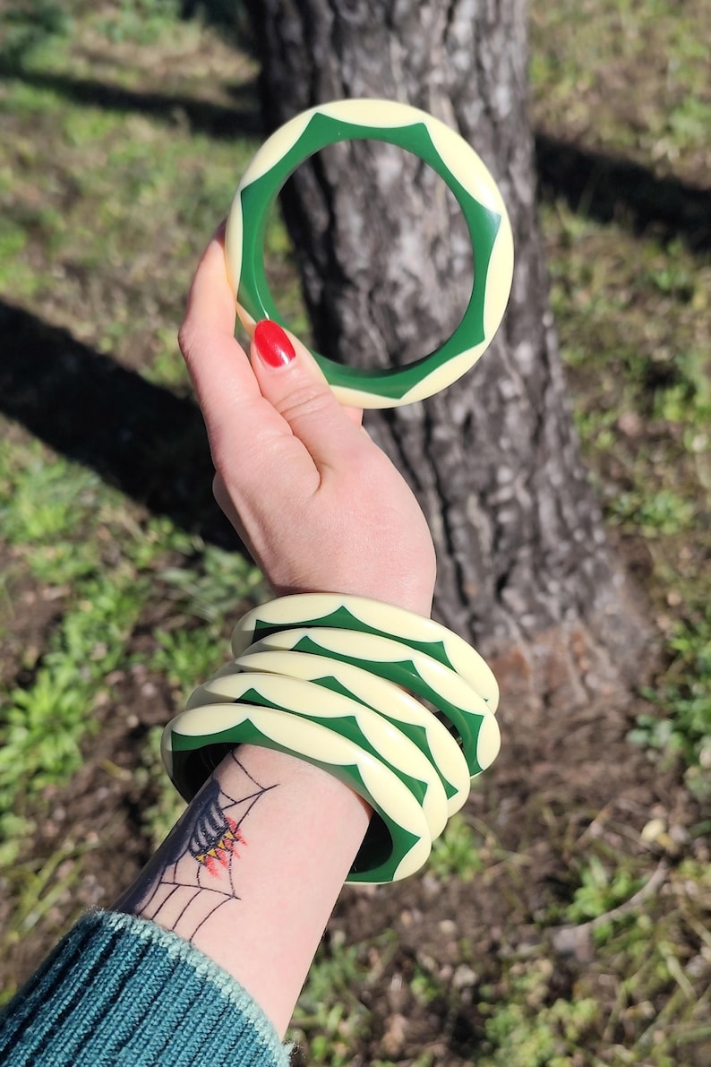 May include: A set of five circular bangle bracelets featuring a repeating pattern of cream-colored and dark green geometric shapes. The bracelets have a smooth, rounded surface and are designed to be worn stacked together on the wrist.