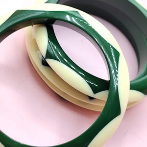 May include: Two circular bangle bracelets featuring a smooth, glossy finish. The design consists of alternating geometric segments in deep forest green and off-white cream colors, creating a repeating pattern around the circumference of each bangle.