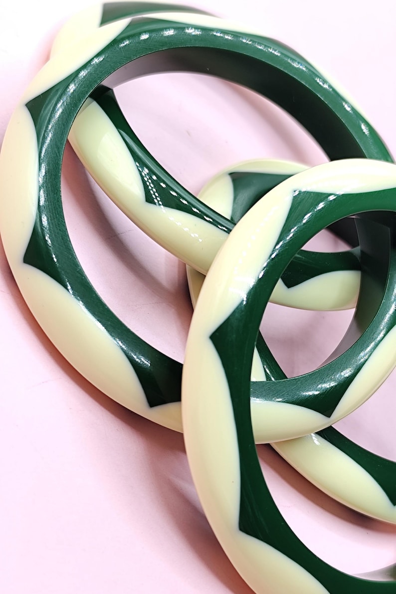 May include: A set of three circular bangles featuring a repeating geometric pattern of alternating dark forest green and cream-colored segments. The smooth, polished surface of each bangle displays a consistent, interlocking chevron-like design.