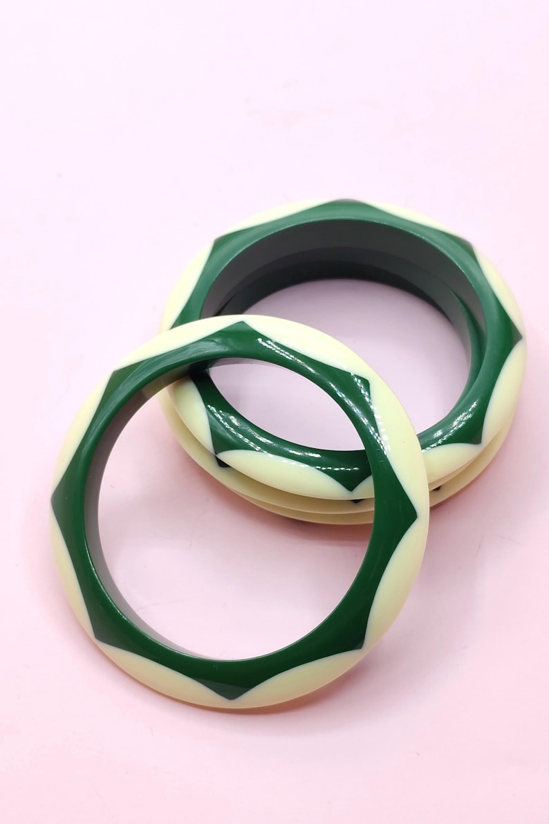 May include: Two circular bangle bracelets featuring a repeating geometric pattern of alternating dark forest green and cream-colored segments. The bracelets have a smooth, polished finish and a thick, rounded profile, creating a bold, high-contrast look.
