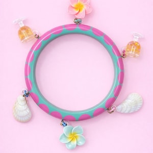 Può includere: Un bracciale rigido circolare con un motivo a pois color verde acqua e rosa. Il bracciale è decorato con cinque ciondoli pendenti: due fiori a cinque petali, uno rosa e uno azzurro con centri gialli, due conchiglie bianche e due piccoli bicchieri trasparenti.