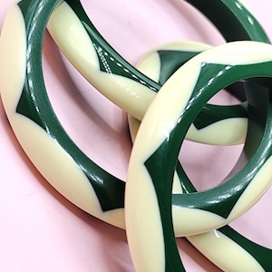 May include: A set of three circular bangles featuring a repeating geometric pattern of alternating dark forest green and cream-colored segments. The smooth, polished surface of each bangle displays a consistent, interlocking chevron-like design.
