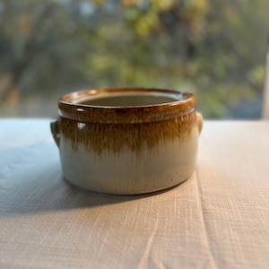 May include: A small, round ceramic bowl with two small handles. The bowl has a white base with a brown, dripped glaze around the rim and handles. The bowl is sitting on a white surface.