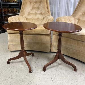 May include: Two matching round side tables made of polished brown wood. Each table features a smooth circular top supported by a central pedestal column that branches into three curved legs at the base, providing a stable foundation.