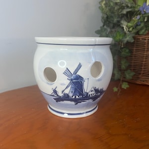 May include: A white and blue ceramic onion keeper with a Dutch windmill design. The round container has three holes and a blue rim and base. The windmill scene is painted in blue, depicting a rural landscape.