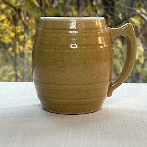 May include: A tan ceramic mug with a handle. The mug has a cylindrical shape with horizontal ridges around the body. The mug is sitting on a white surface, with a blurred background of green foliage.