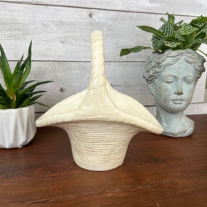 May include: A white ceramic basket with a woven design and a handle. The basket is sitting on a wooden surface.