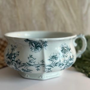 May include: A white ceramic chamber pot with a green floral design. The pot has a single handle and a rounded body.
