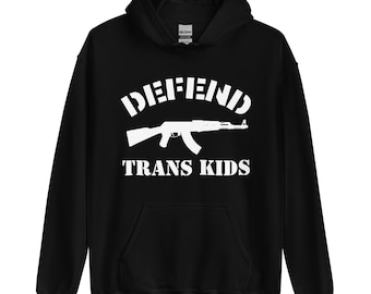 Puede incluir: Una sudadera con capucha negra que presenta un estampado gráfico blanco en el pecho. El diseño incluye el texto DEFEND en una fuente arqueada, una silueta blanca de un rifle tipo AK en el centro y el texto TRANS KIDS debajo del rifle.