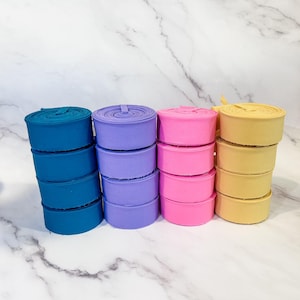 May include: Stacks of colorful fabric rolls in teal, purple, pink, and yellow. Each stack is neatly arranged, showcasing the vibrant hues of the material. The rolls are tightly wound, suggesting a smooth texture, and are ready for crafting or sewing projects.