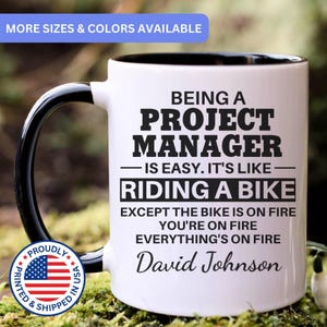 May include: A white ceramic mug with a black interior and handle, featuring black text that reads: BEING A PROJECT MANAGER IS EASY. IT'S LIKE RIDING A BIKE EXCEPT THE BIKE IS ON FIRE YOU'RE ON FIRE EVERYTHING'S ON FIRE David Johnson.