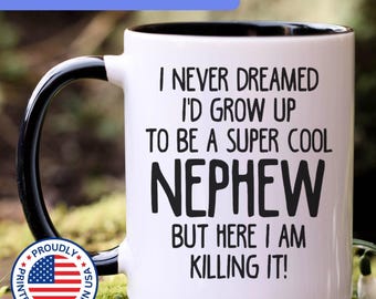 May include: A white ceramic mug with a black interior and handle. The mug features black text that reads: I NEVER DREAMED I'D GROW UP TO BE A SUPER COOL NEPHEW BUT HERE I AM KILLING IT! A circular badge with the American flag and text is also visible.