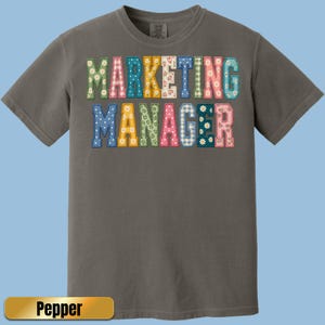May include: A charcoal gray short-sleeve t-shirt featuring the text MARKETING MANAGER in block letters. Each letter has a unique pattern, including floral, gingham, and polka dot prints in various colors like green, pink, blue, and yellow.