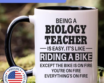May include: A white ceramic mug with a black interior and handle, featuring black text that reads: BEING A BIOLOGY TEACHER IS EASY. IT'S LIKE RIDING A BIKE EXCEPT THE BIKE IS ON FIRE YOU'RE ON FIRE EVERYTHING'S ON FIRE. Includes a circular US flag emblem.
