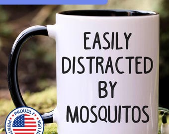 May include: A white ceramic mug with a black handle and interior. The front of the mug features the black text "EASILY DISTRACTED BY MOSQUITOS" in a simple sans-serif font. The mug has a glossy finish and a standard cylindrical shape.