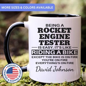 May include: White ceramic mug with a black handle and interior. The mug features the text "BEING A ROCKET ENGINE TESTER IS EASY. IT'S LIKE RIDING A BIKE EXCEPT THE BIKE IS ON FIRE" and the name "David Johnson".