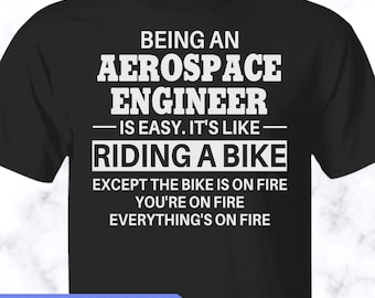 May include: A black short-sleeved t-shirt featuring white text that reads: BEING AN AEROSPACE ENGINEER IS EASY. IT'S LIKE RIDING A BIKE EXCEPT THE BIKE IS ON FIRE YOU'RE ON FIRE EVERYTHING'S ON FIRE. A blue banner at the bottom says MORE STYLES & COLORS AVAILABLE!