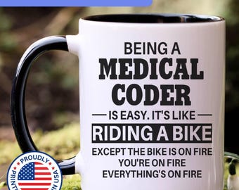 May include: A white ceramic mug with a black interior and handle. The mug features black text that reads: BEING A MEDICAL CODER IS EASY. IT'S LIKE RIDING A BIKE EXCEPT THE BIKE IS ON FIRE YOU'RE ON FIRE EVERYTHING'S ON FIRE. A circular flag emblem is present.
