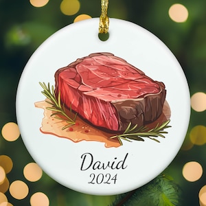 May include: A round white ceramic ornament featuring a colorful illustration of a raw steak garnished with a sprig of green rosemary. Below the steak, the name David and the year 2024 are printed in a black serif font. A gold cord is attached.