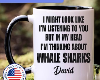 May include: A white ceramic mug with a black interior and handle, featuring the black text: I MIGHT LOOK LIKE I'M LISTENING TO YOU BUT IN MY HEAD I'M THINKING ABOUT WHALE SHARKS, followed by the name David in a black script font.