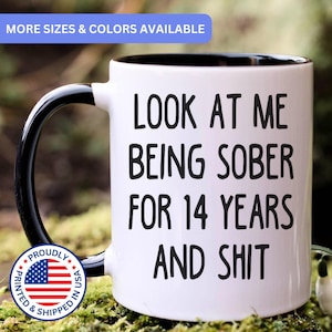 May include: A white ceramic mug with a black interior and handle. The mug features black text that reads: LOOK AT ME BEING SOBER FOR 14 YEARS AND SHIT. A blue banner at the top states MORE SIZES & COLORS AVAILABLE, and a circular badge shows a US flag.