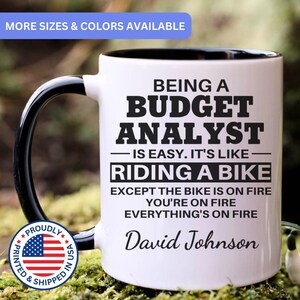 May include: A white ceramic mug with a black interior and handle, featuring black text that reads: BEING A BUDGET ANALYST IS EASY. IT'S LIKE RIDING A BIKE EXCEPT THE BIKE IS ON FIRE YOU'RE ON FIRE EVERYTHING'S ON FIRE. The name David Johnson is printed below.