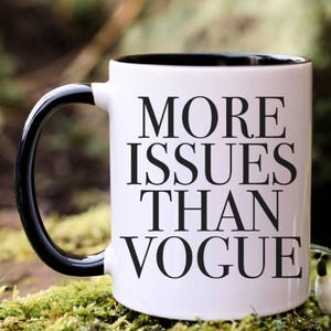 May include: White ceramic mug with a black handle and rim. The mug features the text "MORE ISSUES THAN VOGUE" in bold black letters. The mug is set against a blurred green and brown background.