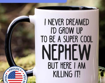 May include: A white ceramic mug with a black interior and handle. The mug features black text that reads: I NEVER DREAMED I'D GROW UP TO BE A SUPER COOL NEPHEW BUT HERE I AM KILLING IT! A circular badge with the American flag and text is also visible.