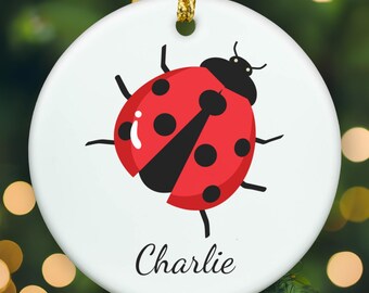 May include: A round white ceramic ornament featuring a red ladybug with black spots and a black head. The name Charlie is printed in black cursive script below the ladybug. A gold string is attached to the top for hanging the ornament.