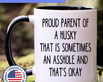 May include: A white ceramic mug with a black interior and handle. The mug features black text that reads: PROUD PARENT OF A HUSKY THAT IS SOMETIMES AN ASSHOLE AND THAT'S OKAY. A circular badge with the American flag and the text PROUDLY PRINTED & SHIPPED IN USA.