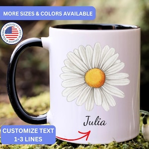 May include: A white ceramic mug with a black handle and interior, featuring a large illustration of a white daisy with a yellow center. The name Julia is printed in black script below the flower. The mug includes text stating MORE SIZES & COLORS AVAILABLE and CUSTOMIZE TEXT 1-3 LINES.