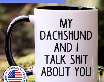 May include: A white ceramic mug with a black interior and a black handle. The front of the mug features the black printed text: MY DACHSHUND AND I TALK SHIT ABOUT YOU. The mug is displayed against a blurred natural background with green moss.